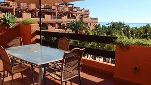 A terrace with a glass table and wicker chairs, overlooking a coastal area with buildings and palm trees. - Ideal and front line beach location (Estepona)