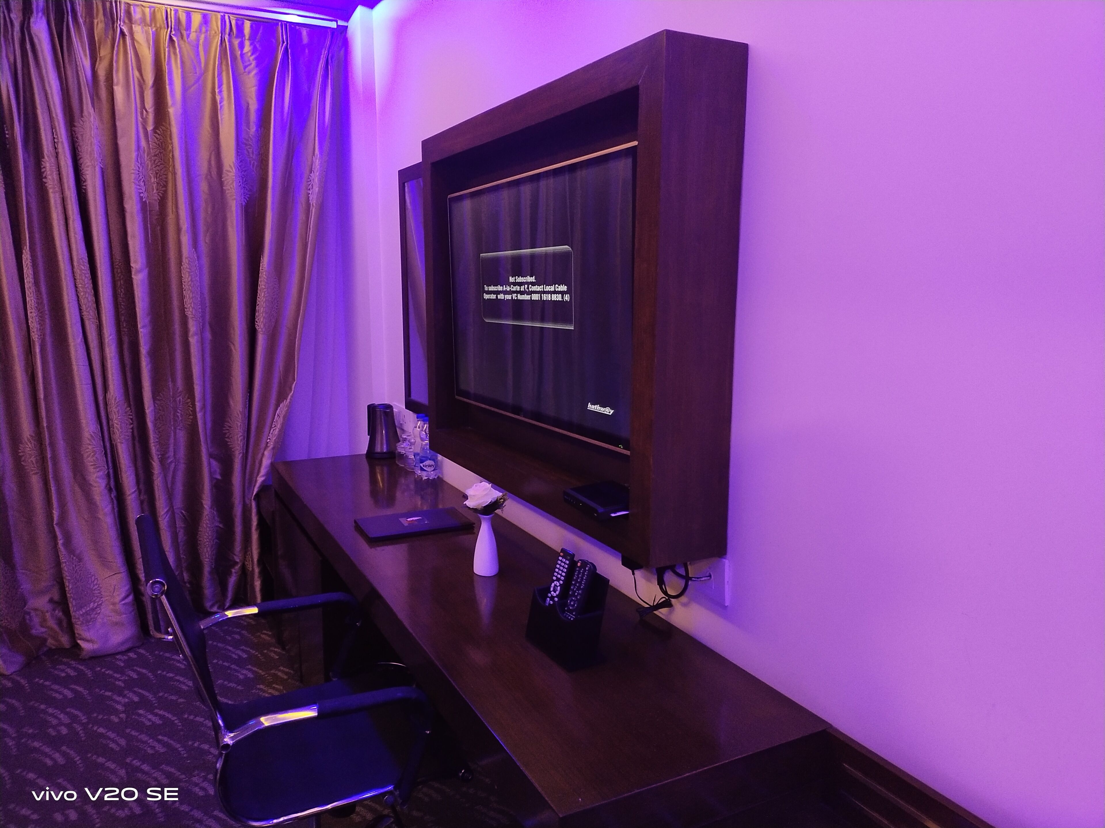 Habitación de hotel con un escritorio de madera oscura, un televisor de pantalla plana en la pared, una silla, una mesita con una botella y una caja de pañuelos, y una cortina con estampado floral.