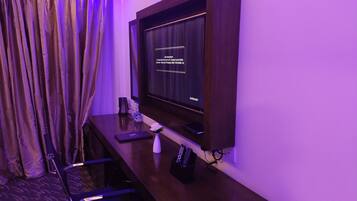 Habitación de hotel con un escritorio de madera oscura, un televisor de pantalla plana en la pared, una silla, una mesita con una botella y una caja de pañuelos, y una cortina con estampado floral.