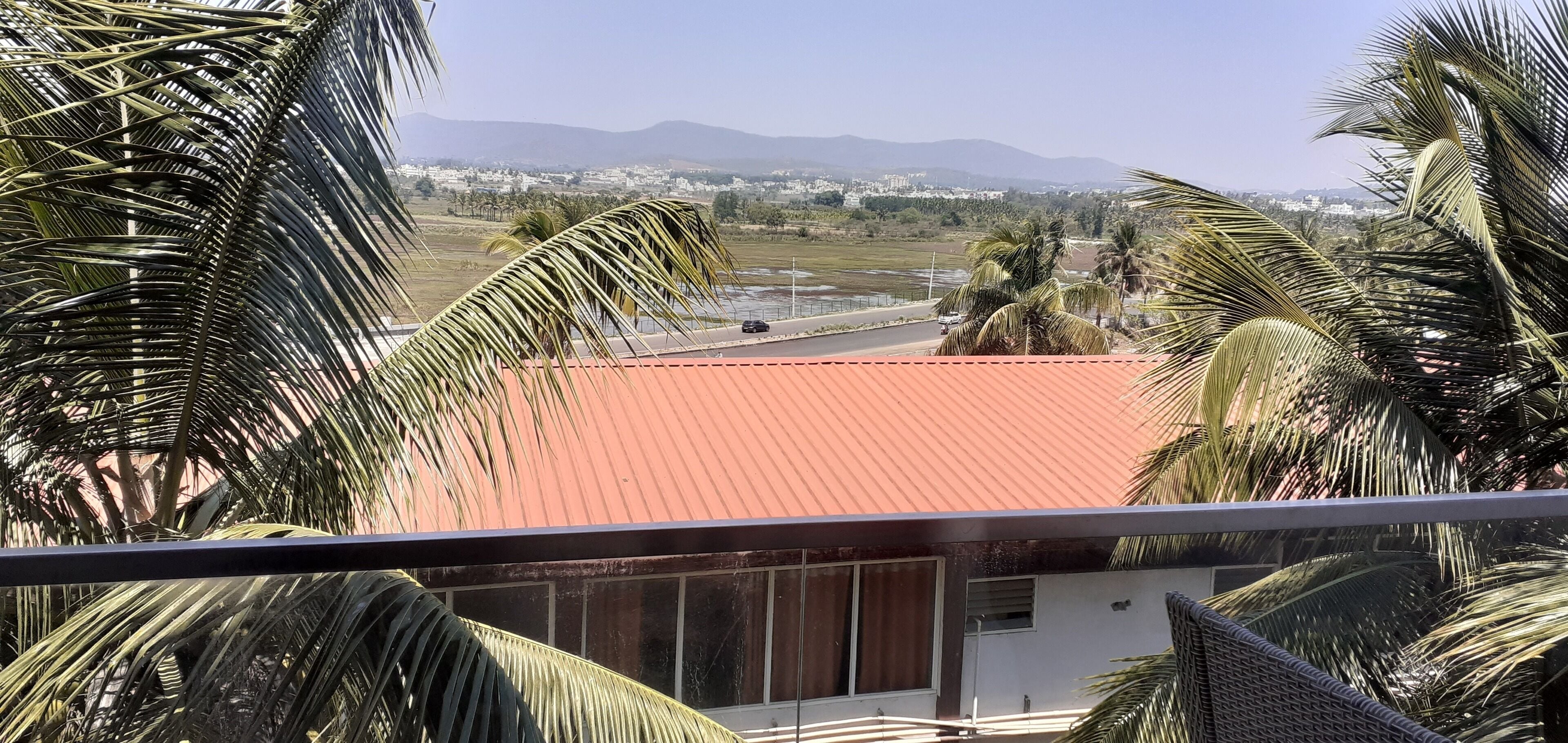 Vista desde un balcón con techo rojo, palmeras y un paisaje distante con edificios y montañas.