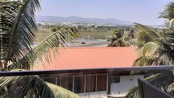 Vista desde un balcón con techo rojo, palmeras y un paisaje distante con edificios y montañas.