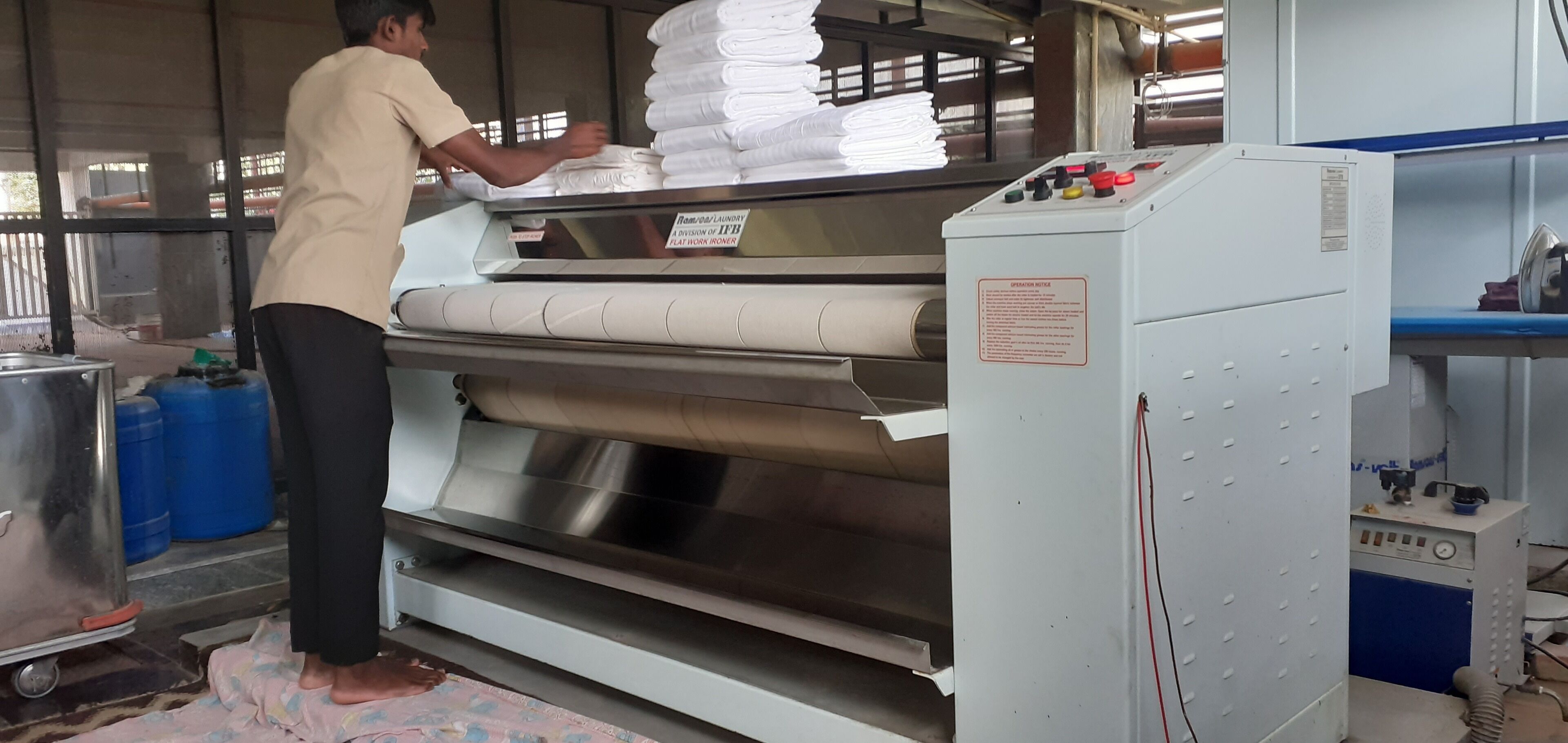 Una persona opera una máquina industrial grande con un panel de control, rodeada de pilas de toallas o ropa blanca.
