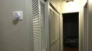 Un couloir avec un thermostat, des volets blancs et un lit recouvert d’une literie à motifs.