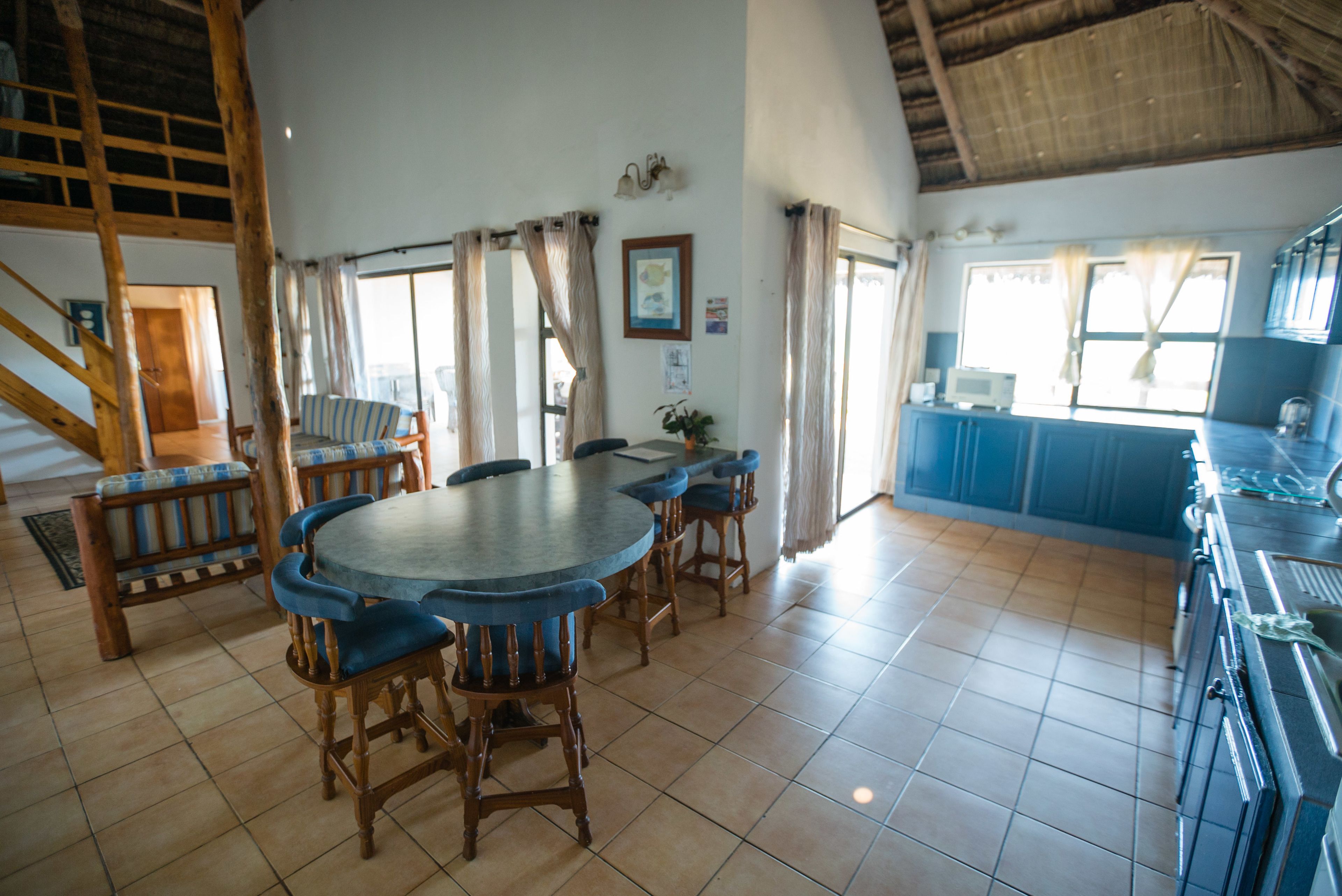 A spacious dining area with a large round table and six chairs, a kitchen area with blue cabinets, and a staircase leading to an upper level.
