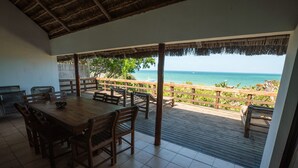 Een overdekt terras met een houten tafel en stoelen, een rieten dak en uitzicht op de oceaan.