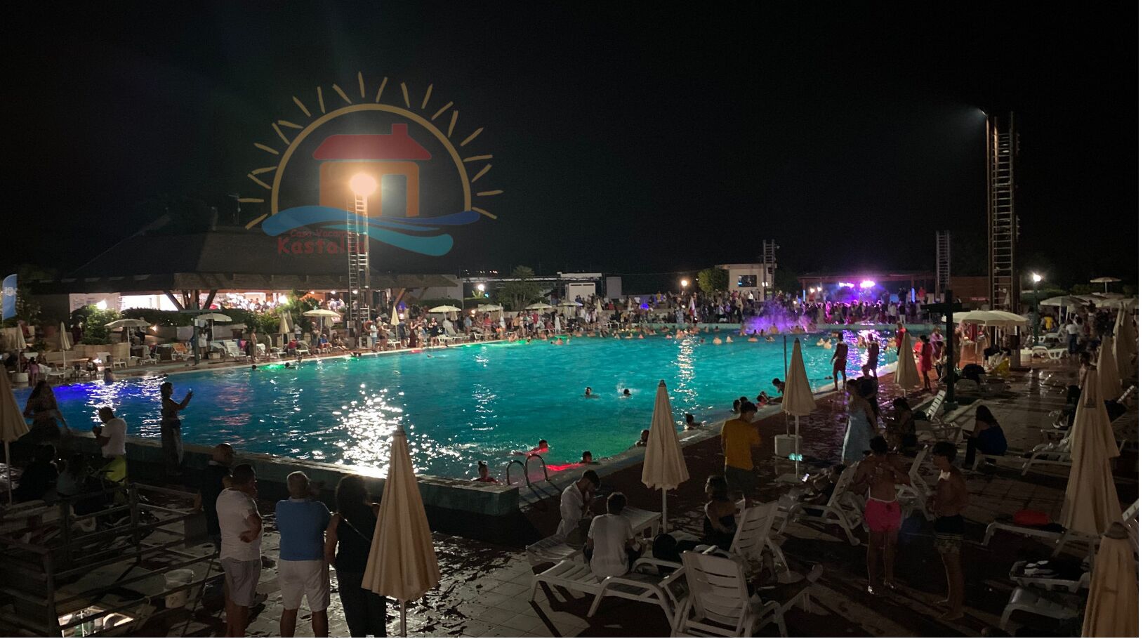 Ein Poolbereich bei Nacht mit Liegestühlen und Sonnenschirmen, eine große beleuchtete Schautafel und viele Menschen.