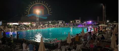Ein Poolbereich bei Nacht mit Liegestühlen und Sonnenschirmen, eine große beleuchtete Schautafel und viele Menschen.