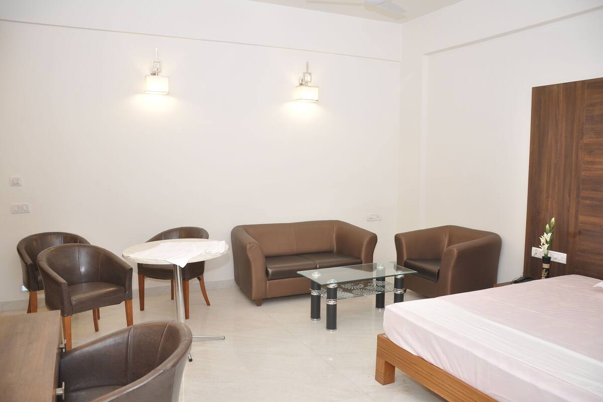 Una habitación de hotel moderna con un sofá de cuero marrón, dos sillones, una mesita de vidrio, una pequeña mesa redonda y una cama con colcha blanca.