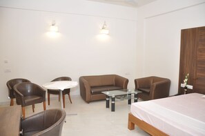 A modern hotel room with a brown leather sofa, two armchairs, a glass coffee table, a small round table, and a bed with a white bedspread.