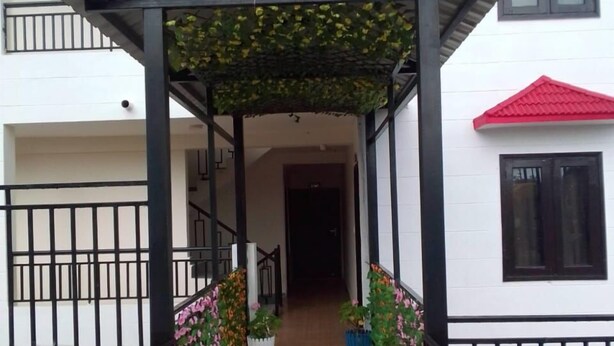 A covered walkway with potted plants, leading to a building with a red roof and white walls. - S L Residency (Dhari)