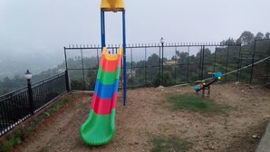 A playground with a slide and swings, surrounded by a fence and a view of distant hills. - S L Residency (Dhari)