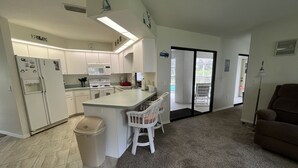 Eine kompakte Küche mit Kühlschrank, Herd und Mikrowelle. Es gibt eine kleine Essecke mit einem Stuhl und einem Mülleimer. Der Raum hat einen Teppichboden und eine Schiebetür, die auf einen Balkon führt.