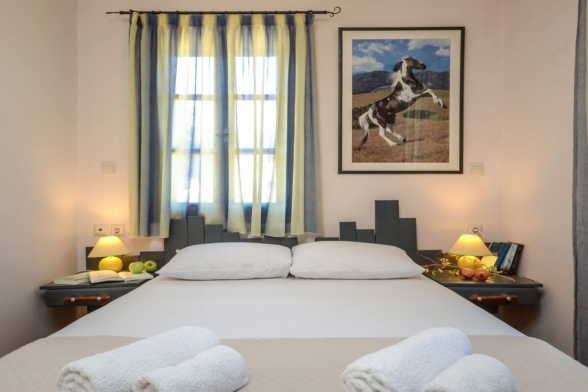 Quarto de hotel com uma cama grande, duas mesas de cabeceira com luminárias, uma janela com cortinas e um quadro pendurado na parede.