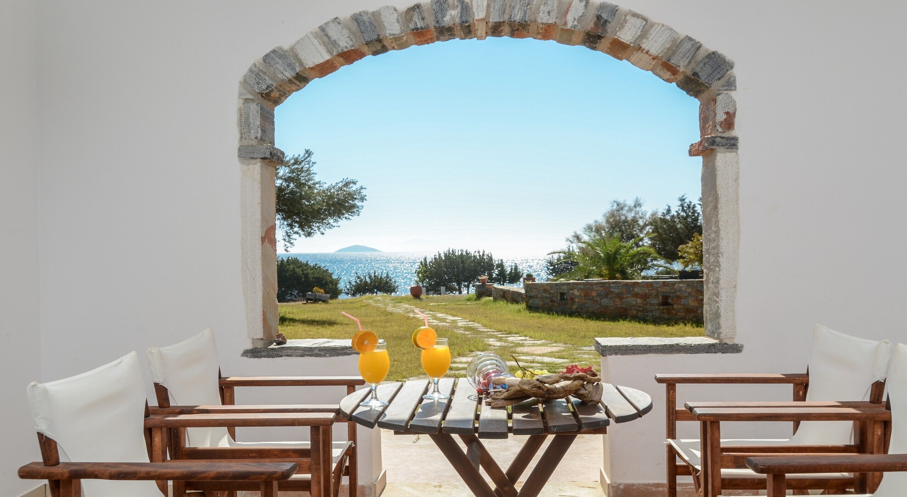 A table with two chairs, a wooden table, and a view through an archway to a grassy area with a stone wall and trees.