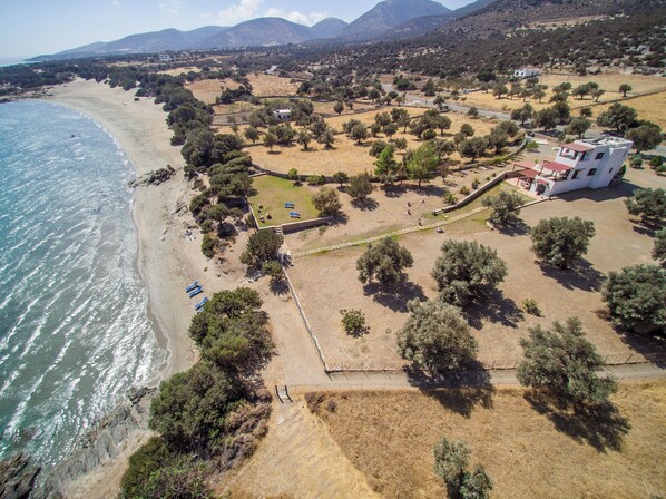 Vue aérienne d’une zone côtière comprenant une plage, une propriété clôturée avec un bâtiment et quelques arbres dispersés.
