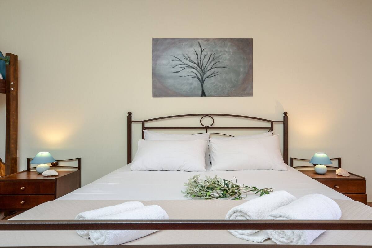 Uma cama bem arrumada com lençóis brancos, duas luminárias de cabeceira, uma pintura de uma árvore e uma pequena planta na cama.