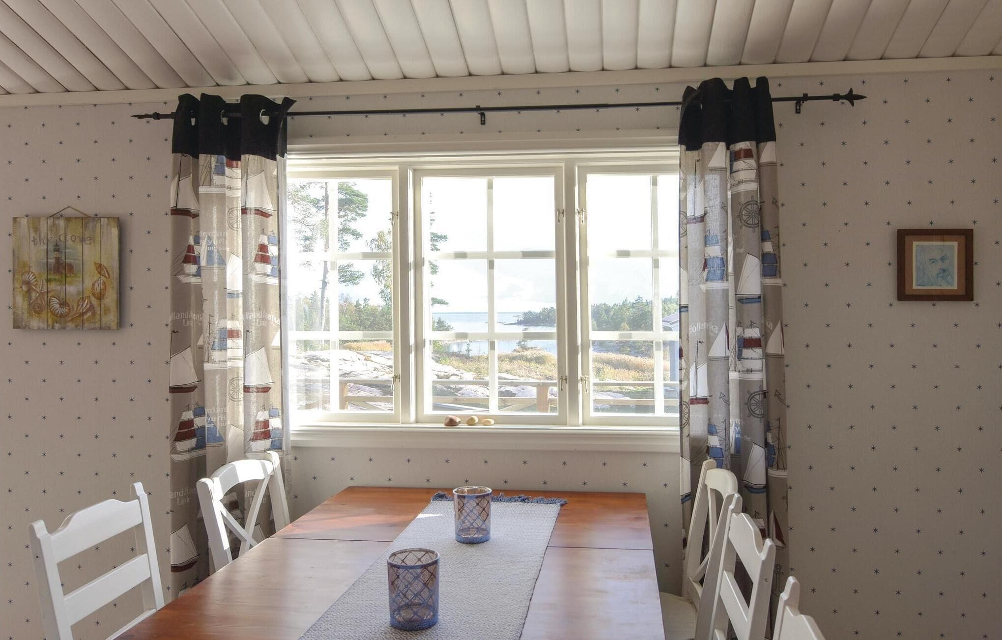 A dining area with a wooden table, white chairs, a window with curtains, and a view of a landscape.