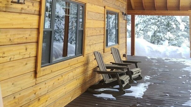 Een houten blokhut met een overdekte veranda, grote ramen en uitzicht op besneeuwde bomen.