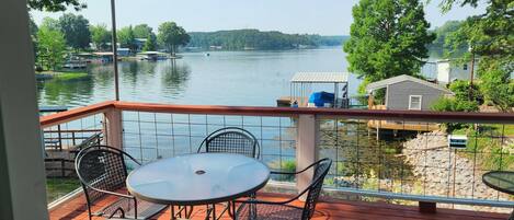 A deck with a table and chairs overlooking a lake, with boats and buildings in the background.
