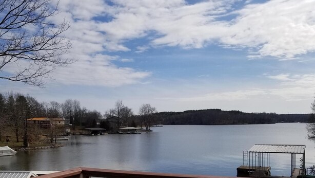 A wooden deck with a metal railing overlooking a calm lake, surrounded by trees and buildings.