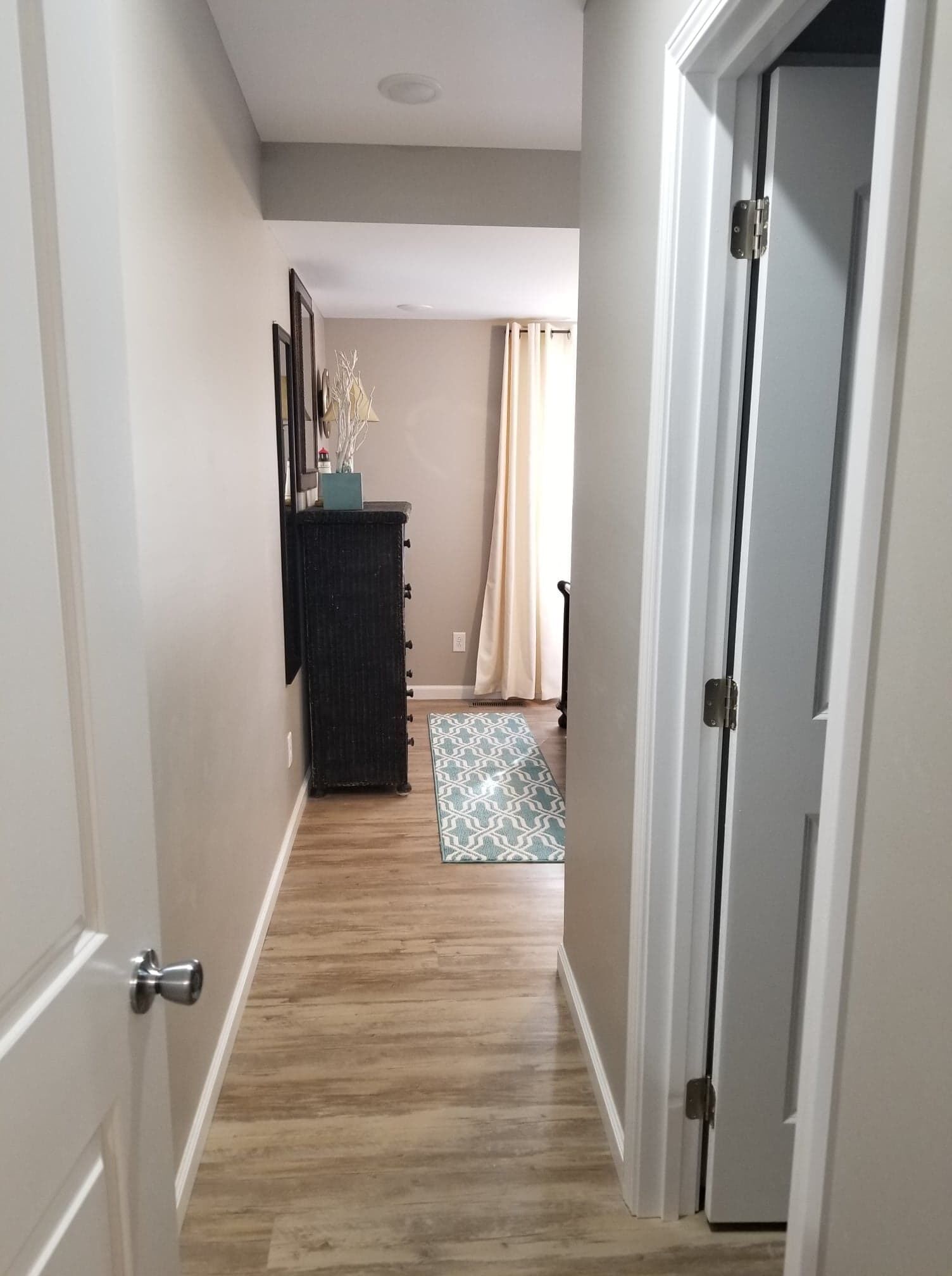 A narrow hallway with wooden flooring, a black cabinet, a patterned rug, and a door with a metal handle.