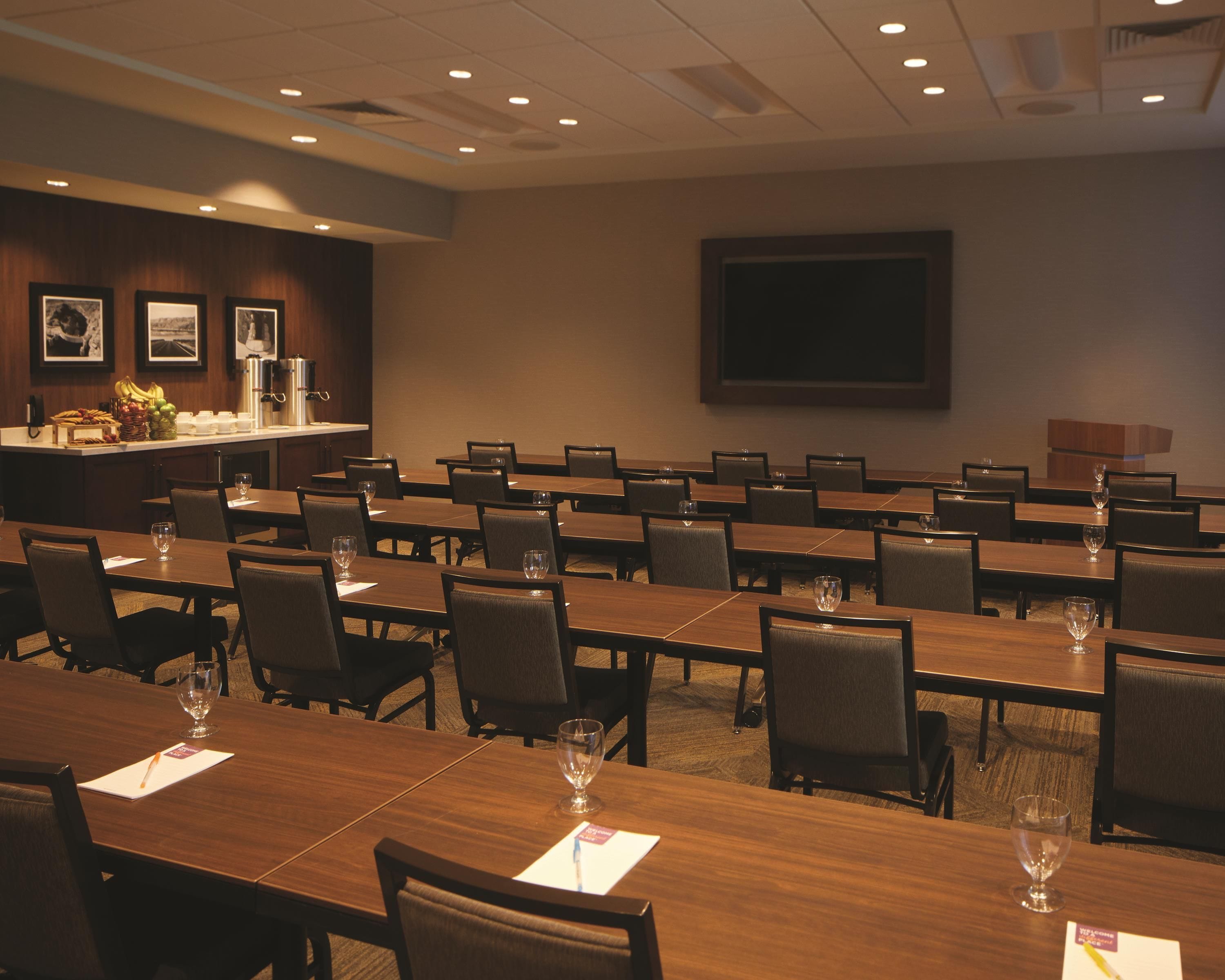 A conference room with multiple tables and chairs, a buffet area with fruits and beverages, and a large screen on the wall.