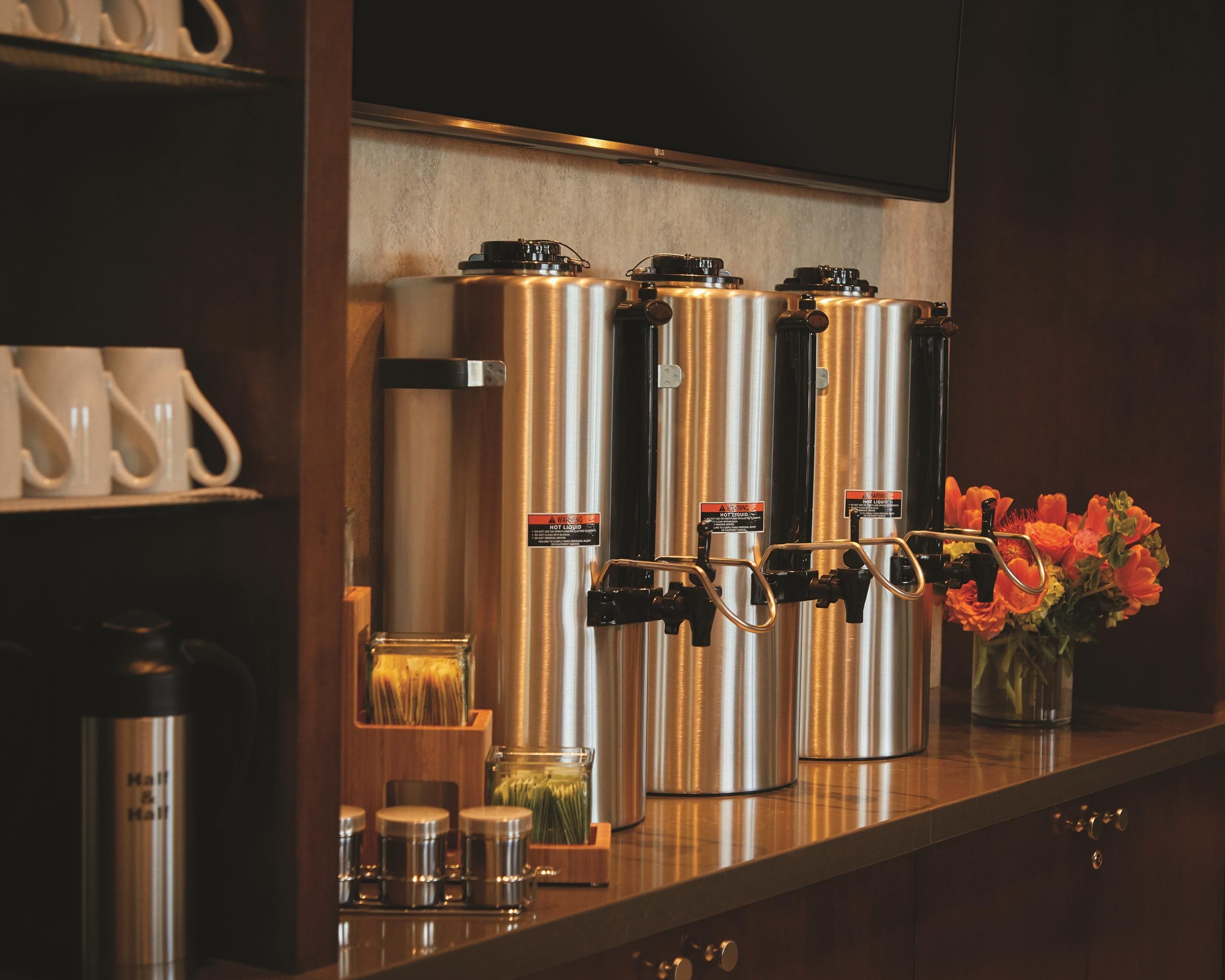 A coffee station with four stainless steel dispensers, a vase with flowers, and a shelf with cups and a thermos.