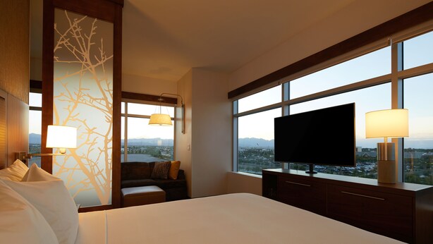 Une chambre d’hôtel dotée d’un grand lit, d’un téléviseur à écran plat, d’un bureau avec une lampe, offrant une vue sur la ville grâce à de grandes fenêtres, et dont le mur est décoré d’un motif représentant des arbres.
