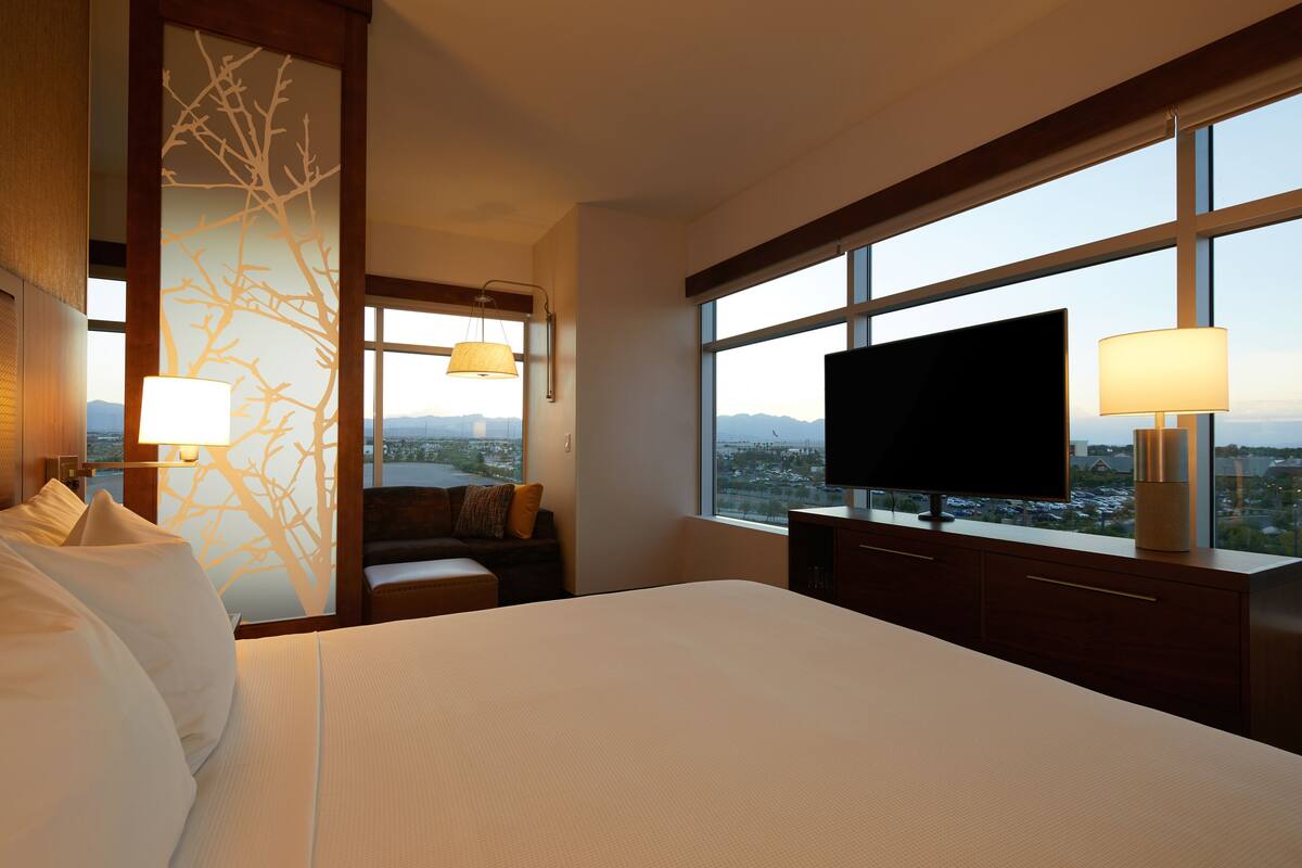 Habitació d'hotel amb un llit gran, televisió de pantalla plana, escriptori amb làmpada, vista de la ciutat a través de grans finestres i un dibuix decoratiu d'un arbre a la paret.