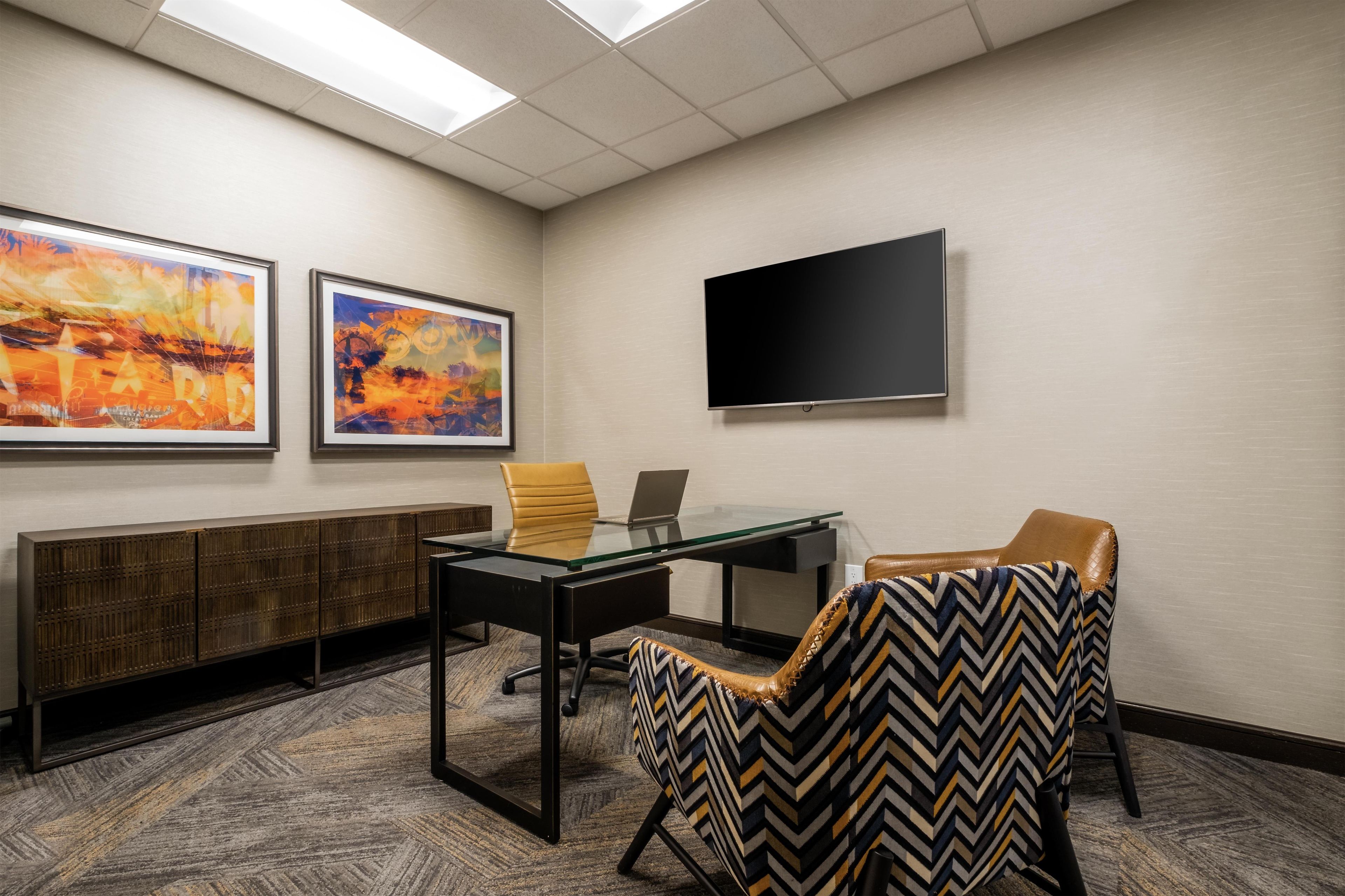 A conference room with a glass table, two chairs, a flat-screen TV, and two framed paintings on the wall.