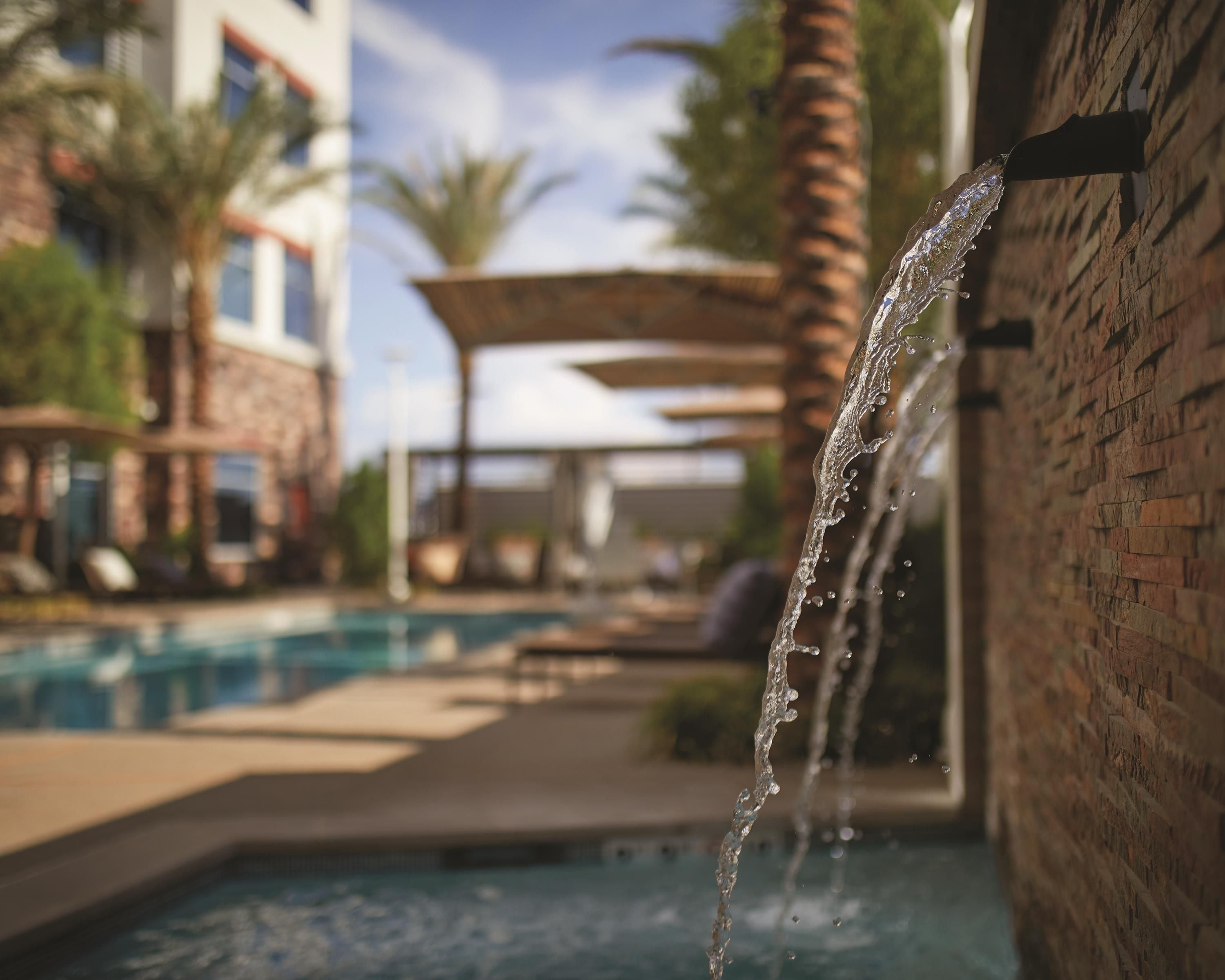 A water feature with a clear stream flowing over a brick wall, a pool, and lounge chairs in the background.
