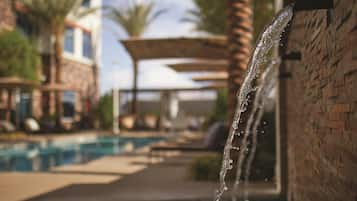A water feature with a clear stream flowing over a brick wall, a pool, and lounge chairs in the background.