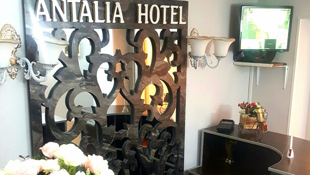 A reception area with a decorative sign reading "ANTALIA HOTEL," a reception desk, a television mounted on the wall, and a vase with flowers on the desk. - Hotel Antalia (Tbilisi)