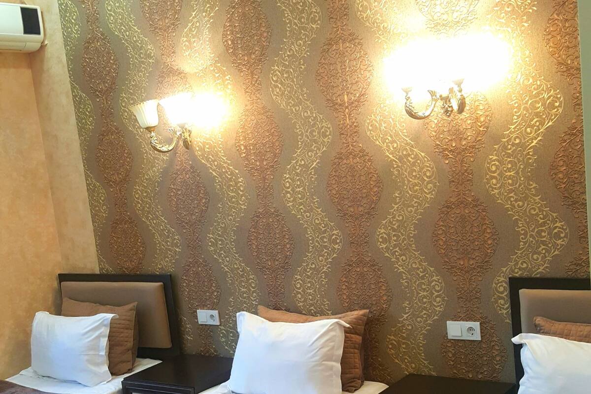 A hotel room with three beds, each with a brown quilted bedspread and white pillows. There are wall sconces with lights on, and a decorative gold-patterned wallpaper.