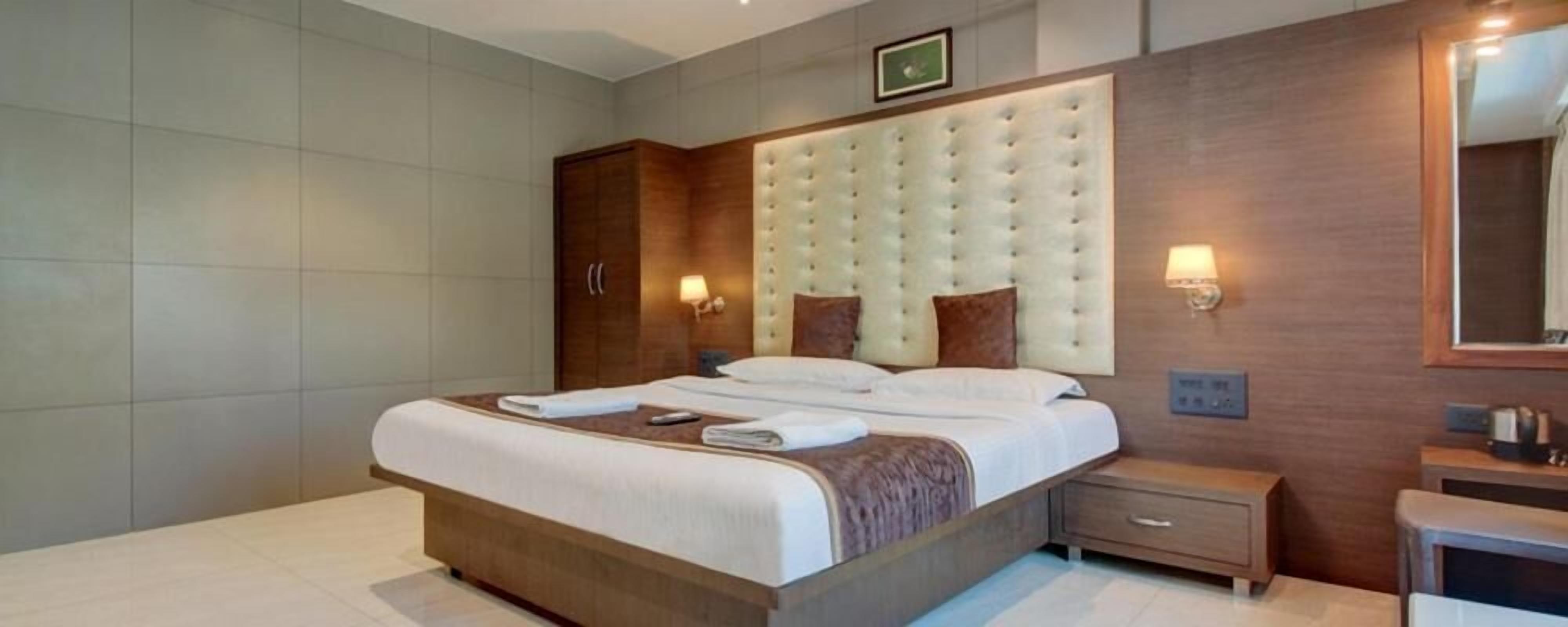 A modern hotel room with a large bed, wooden headboard, bedside tables, and a mirror.