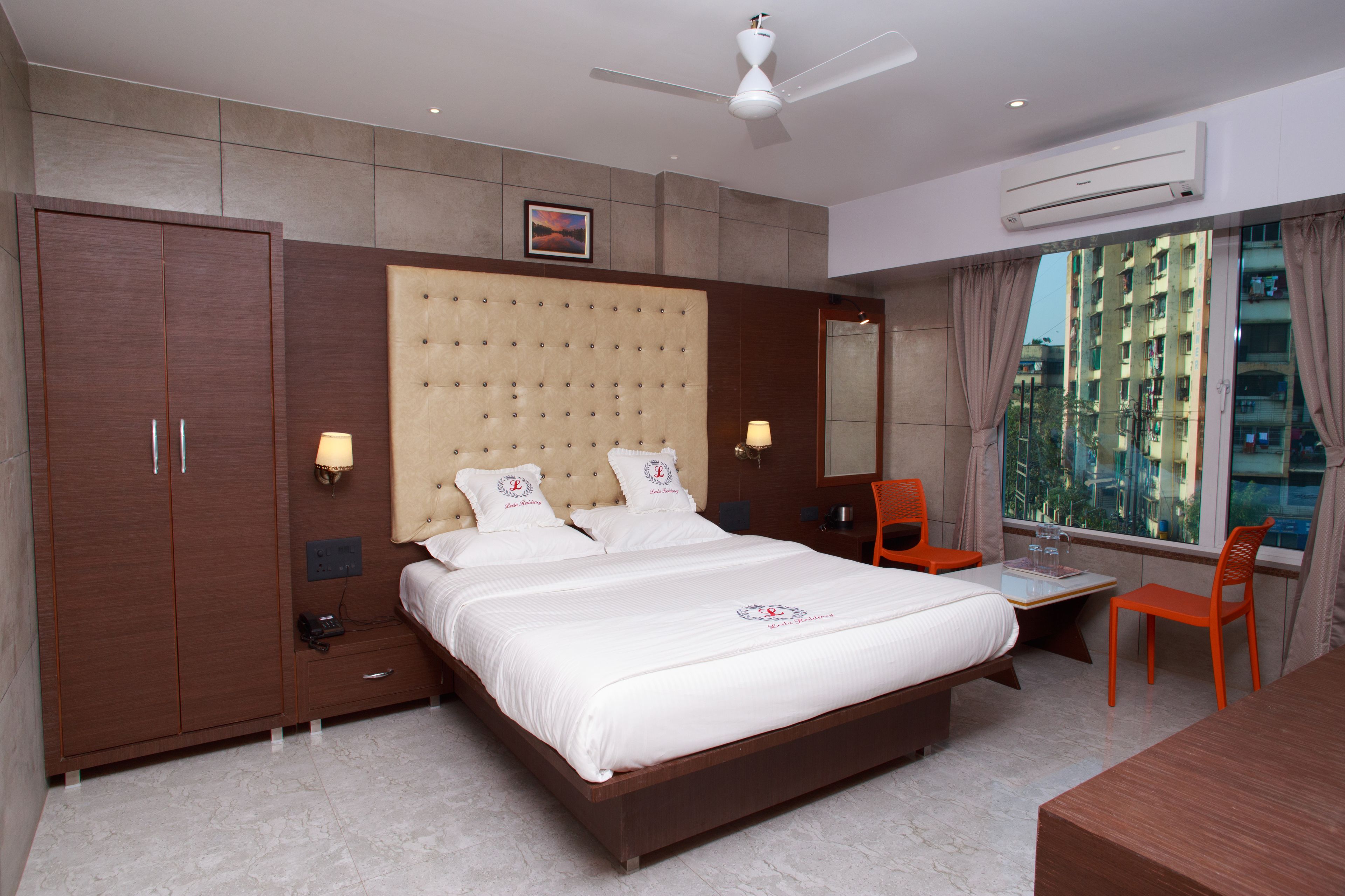 A hotel room with a large bed, a desk, a chair, a wardrobe, and a window with a view of buildings.