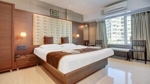 A hotel room with a large bed, a wooden wardrobe, a bedside table, a desk, a chair, a window with curtains, and an air conditioning unit. - Hotel Leela (Kalyan)