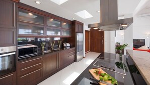 A modern kitchen with dark wood cabinets, a stainless steel refrigerator, a microwave, and a large island with a cutting board and vegetables. - Sunny Banks - 4 Bed Private Pool Sea View Villa (Ko Samui)