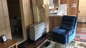 Una habitación con detalles de madera, un sillón azul, un calefactor blanco, una zona de cocina con refrigerador y una alfombra estampada.