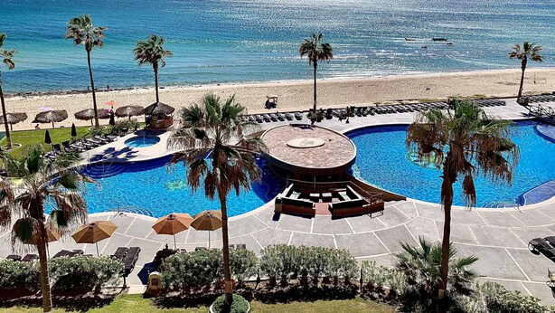 Un espace piscine aménagé dans un complexe hôtelier, avec une fontaine circulaire, des chaises longues, des palmiers et une vue sur la plage.