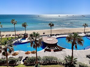 A resort pool area with a circular water feature, lounge chairs, palm trees, and a beach view.