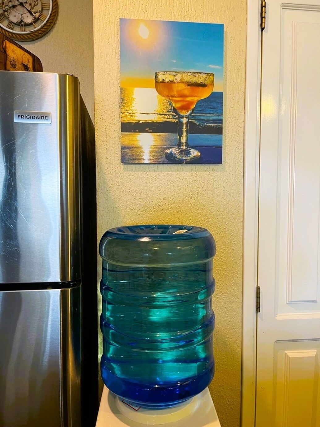 A kitchen with a stainless steel refrigerator, a water dispenser, and a painting of a sunset with a margarita.
