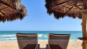 Deux chaises de plage, placées sous des parasols en chaume, sont tournées vers l’océan.