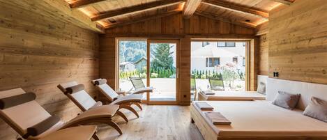 A wooden cabin interior with a bed, lounge chairs, and a large window.