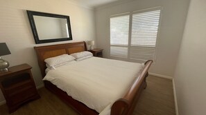 A bedroom with a wooden bed, a nightstand, a lamp, a mirror, and a window with blinds.