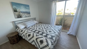 Una camera da letto con un letto, uno sgabello di legno, un quadro appeso al muro e vista sull'esterno attraverso una finestra.