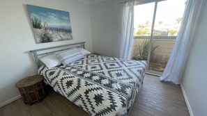 Ein Schlafzimmer mit einem Bett, einem kleinen Holzschemel, einem Bild an der Wand und Blick ins Freie durch ein Fenster.