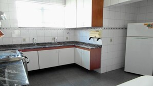 Une cuisine avec des meubles blancs, un plan de travail en granit, un réfrigérateur blanc et une fenêtre ornée d’une croix sur le mur.