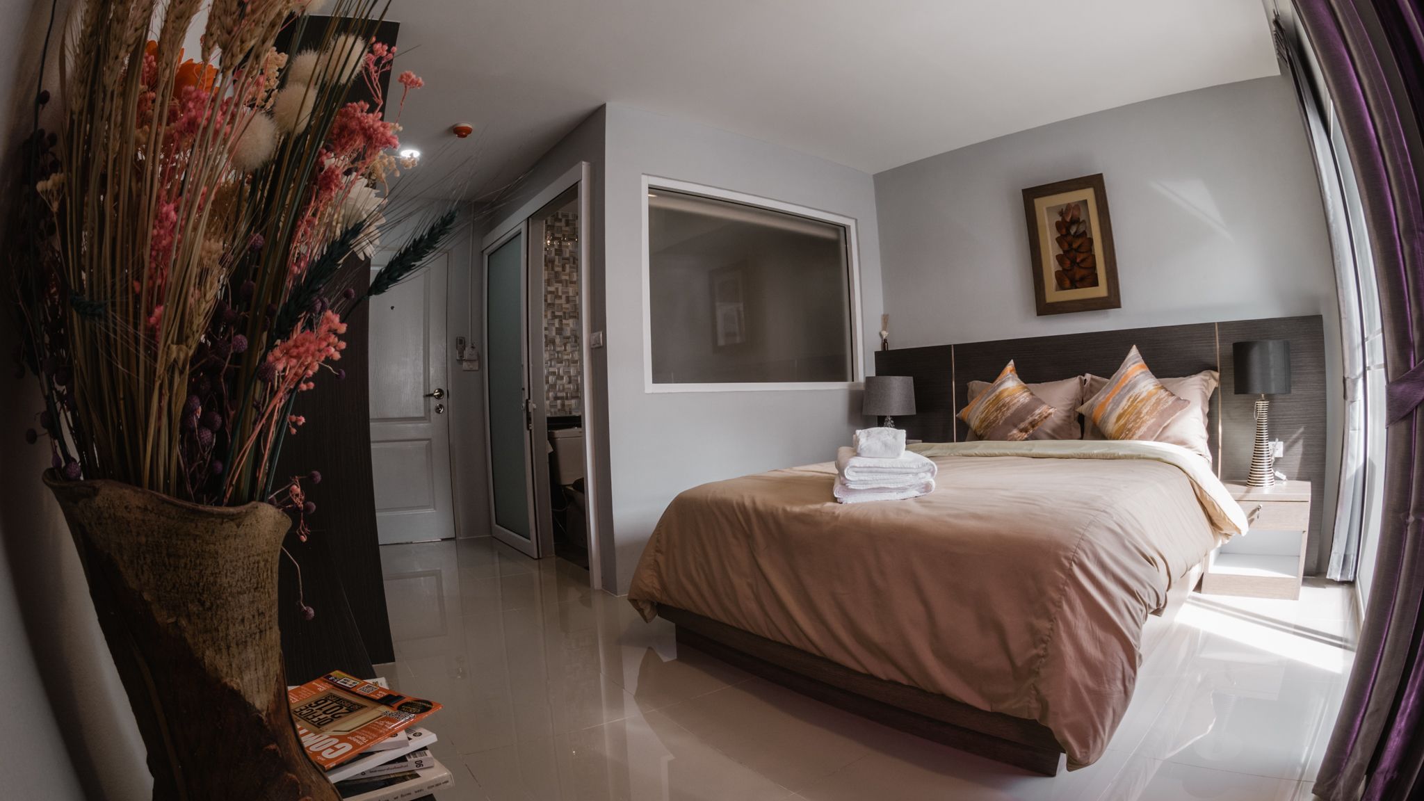 A modern hotel room with a large bed, a bedside table with a lamp, and a view of the hallway.
