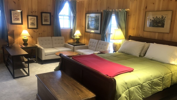 Une chambre d’hôtel avec un mur lambrissé en bois, un lit recouvert d’une courtepointe verte, deux fauteuils blancs, une table de chevet en bois avec une lampe et des œuvres d’art encadrées accrochées au mur.